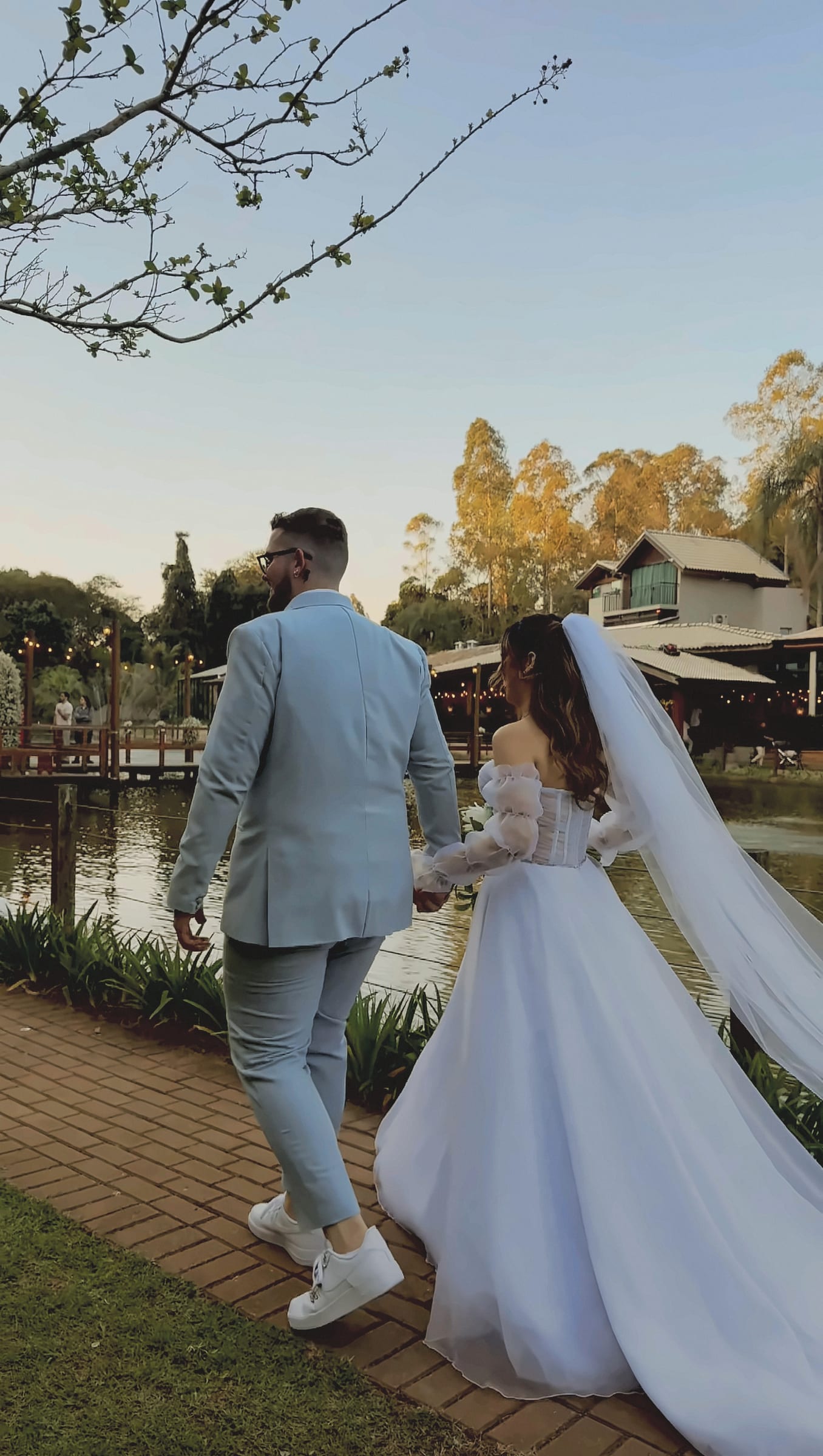 Leonardo e Graziela de mãos dadas caminhando à beira do lago ao entardecer, ele de terno claro e tênis branco, ela de vestido branco com véu longo, o deck e a casa do lago ao fundo.