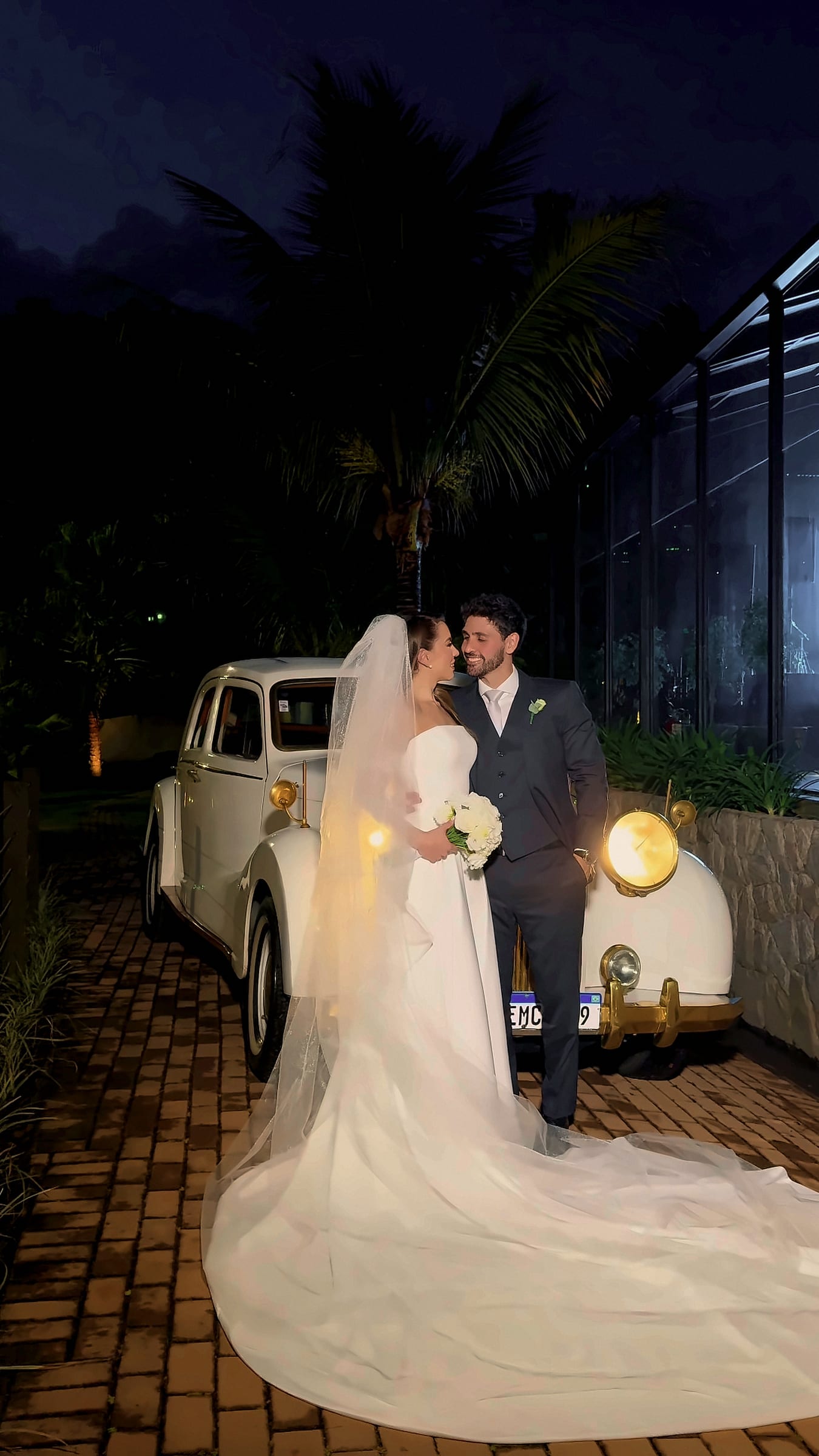 Paola e Gabriel à noite ao lado de um carro antigo branco diante da casa de vidro, ela de vestido branco com véu longo e buquê, ele de terno escuro.