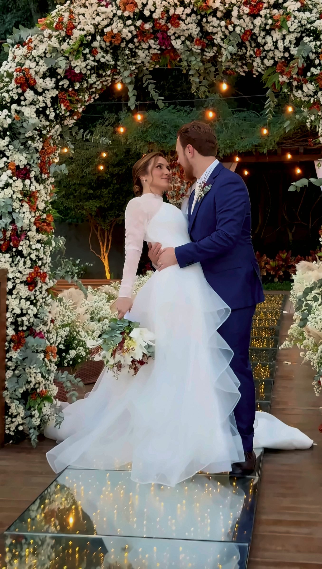Cristiane e Yurguen abraçados sob o arco de flores brancas e vermelhas do deck, com as luzes acesas, ela de vestido branco de mangas de tule, ele de terno azul.