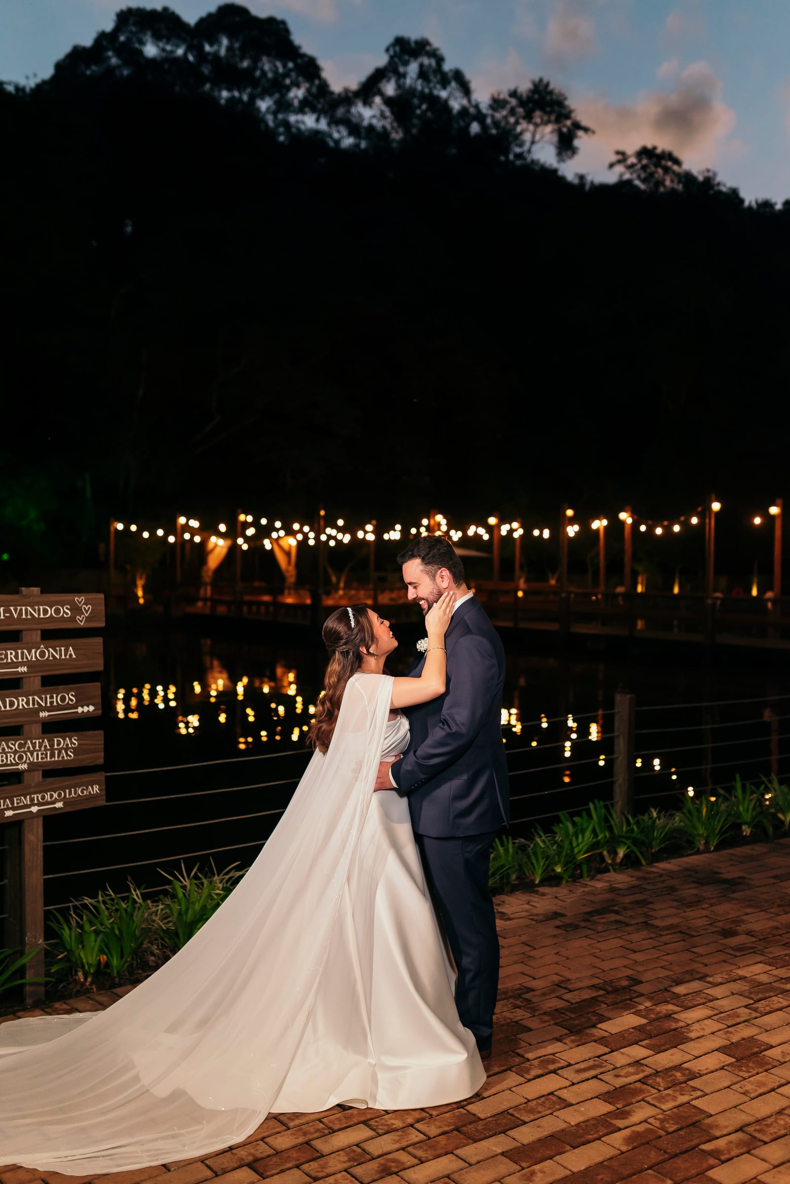 Vinícius e Luciana à beira do lago ao anoitecer, as luzes do deck refletidas na água, ela de vestido branco com véu longo tocando o rosto dele, ele de terno escuro.