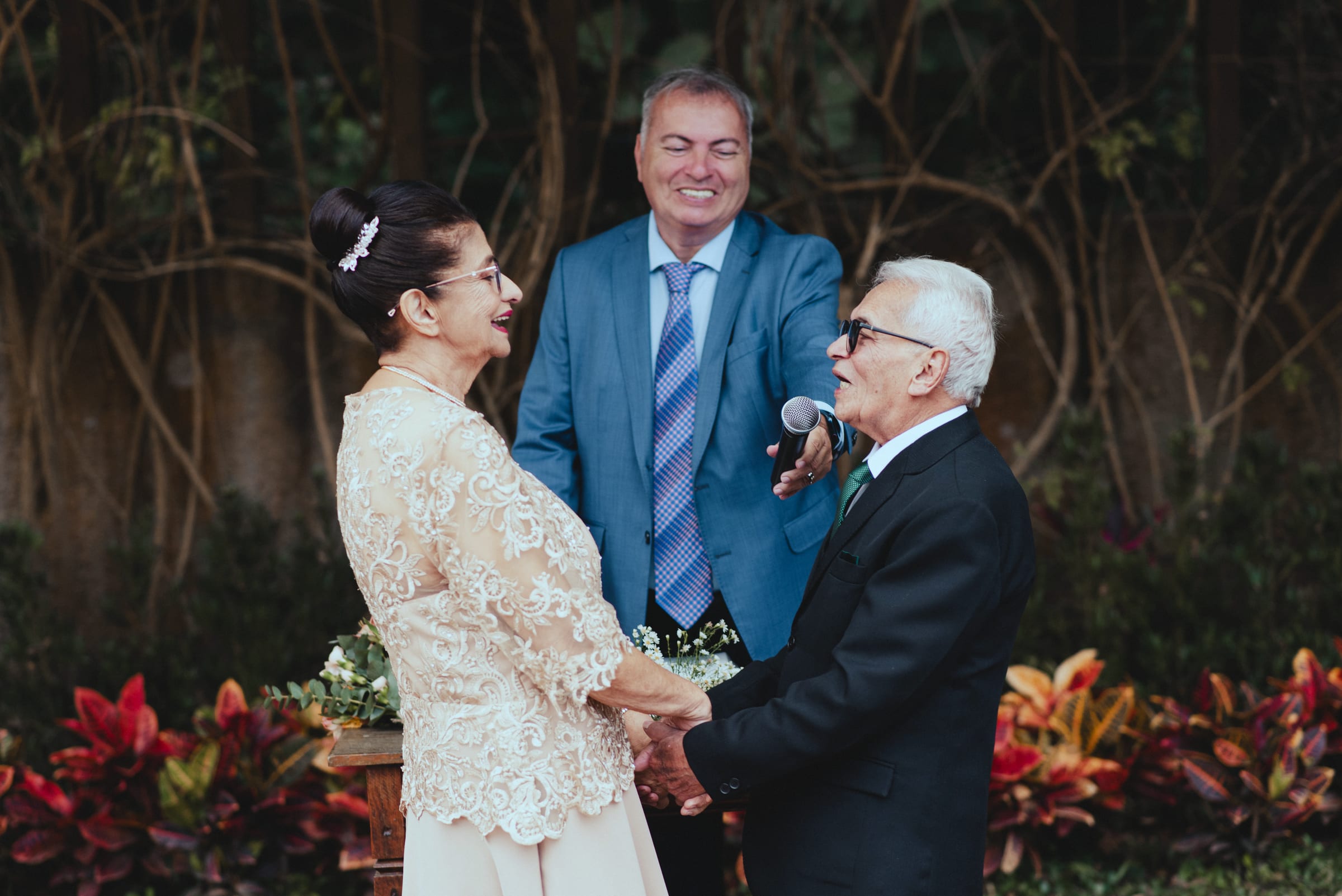 Casal de bodas de mãos dadas diante do celebrante ao microfone, entre as folhagens do jardim.
