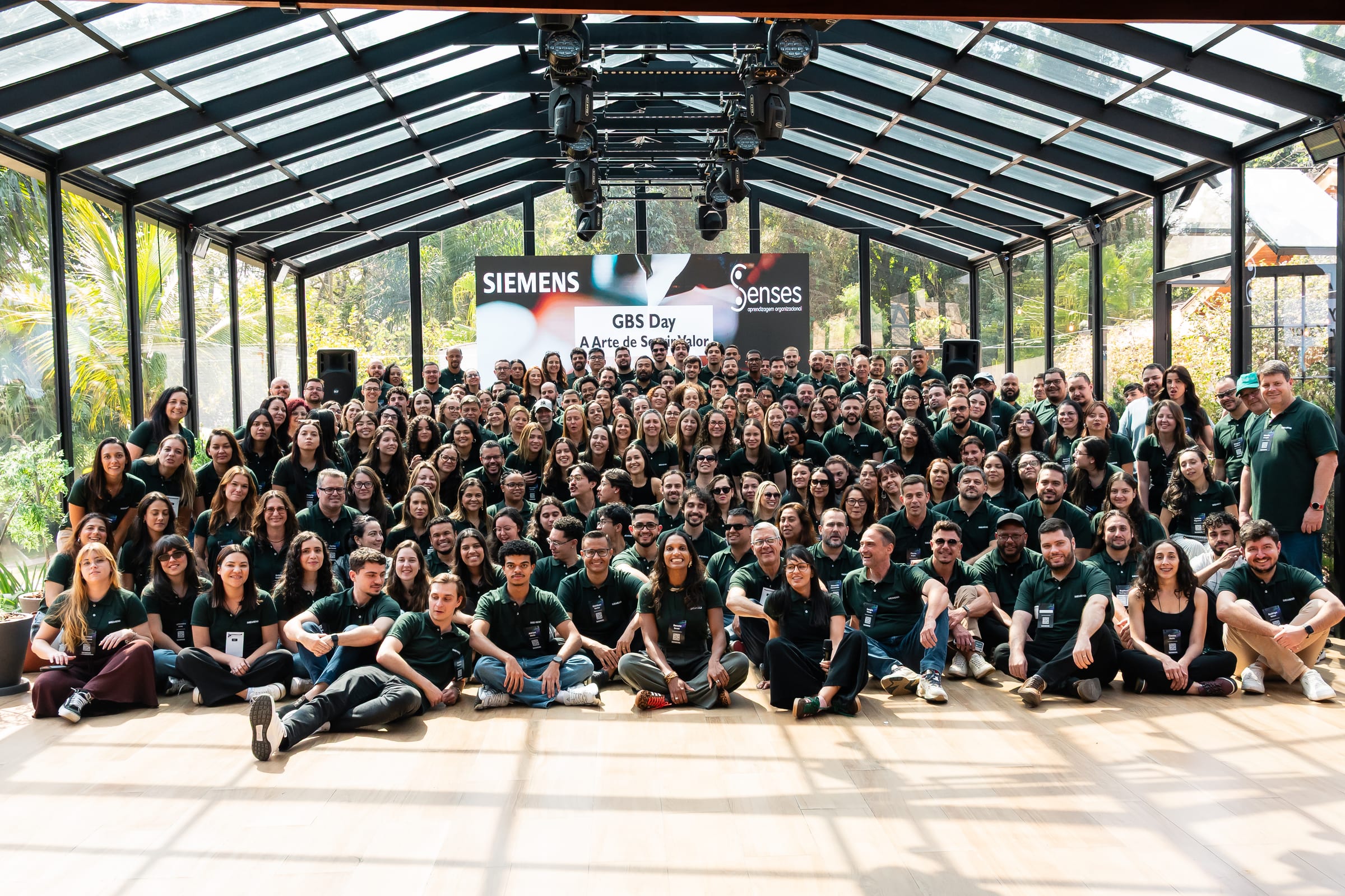 A equipe de uma empresa reunida para a foto sob o telhado da casa de vidro, com o painel do evento ao fundo.