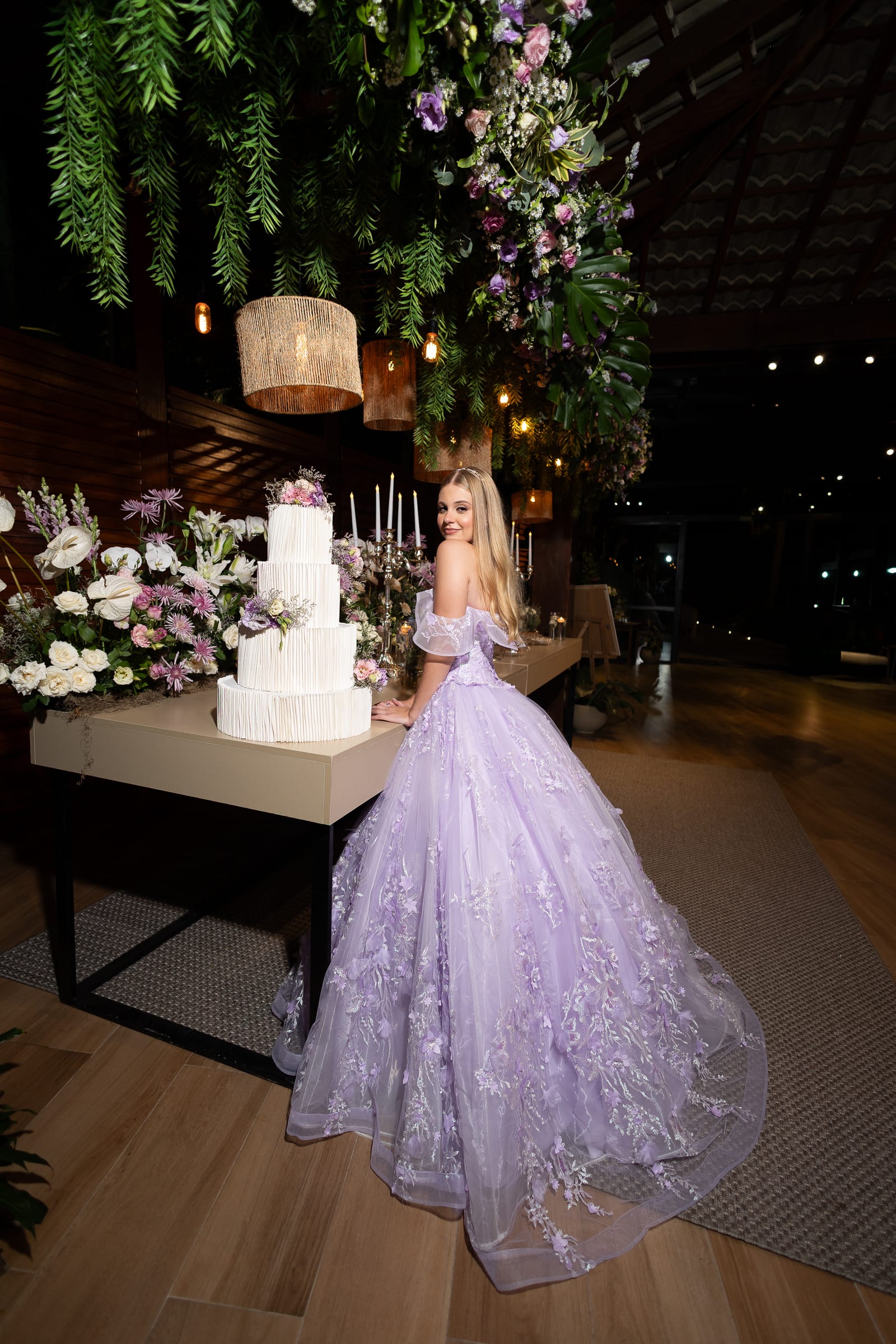 Debutante de vestido lilás rodado ao lado da mesa do bolo, sob as flores suspensas do salão.