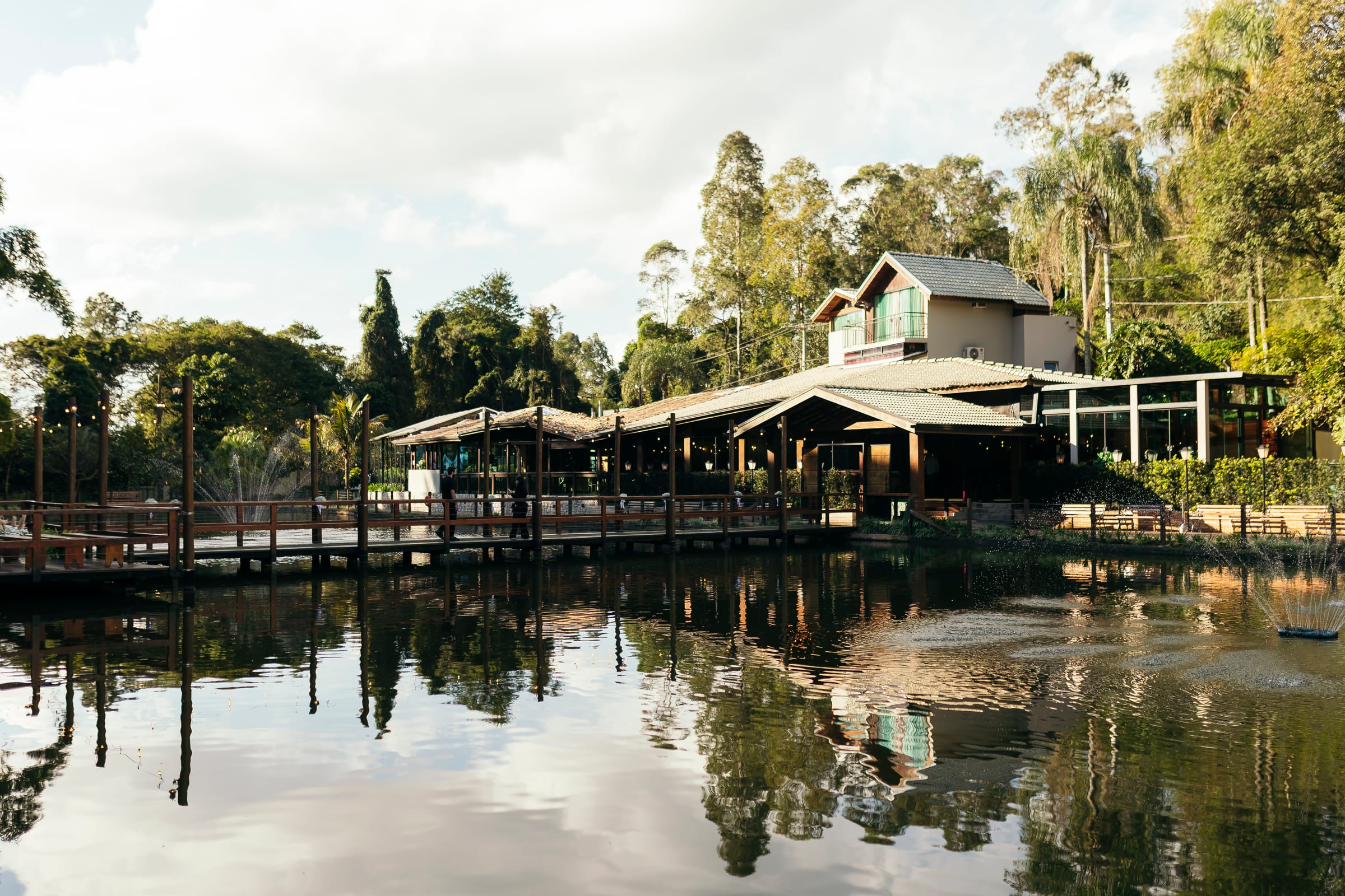 O lago do Acqua com o salão e o deck refletidos na água parada, a mata e o céu claro atrás.