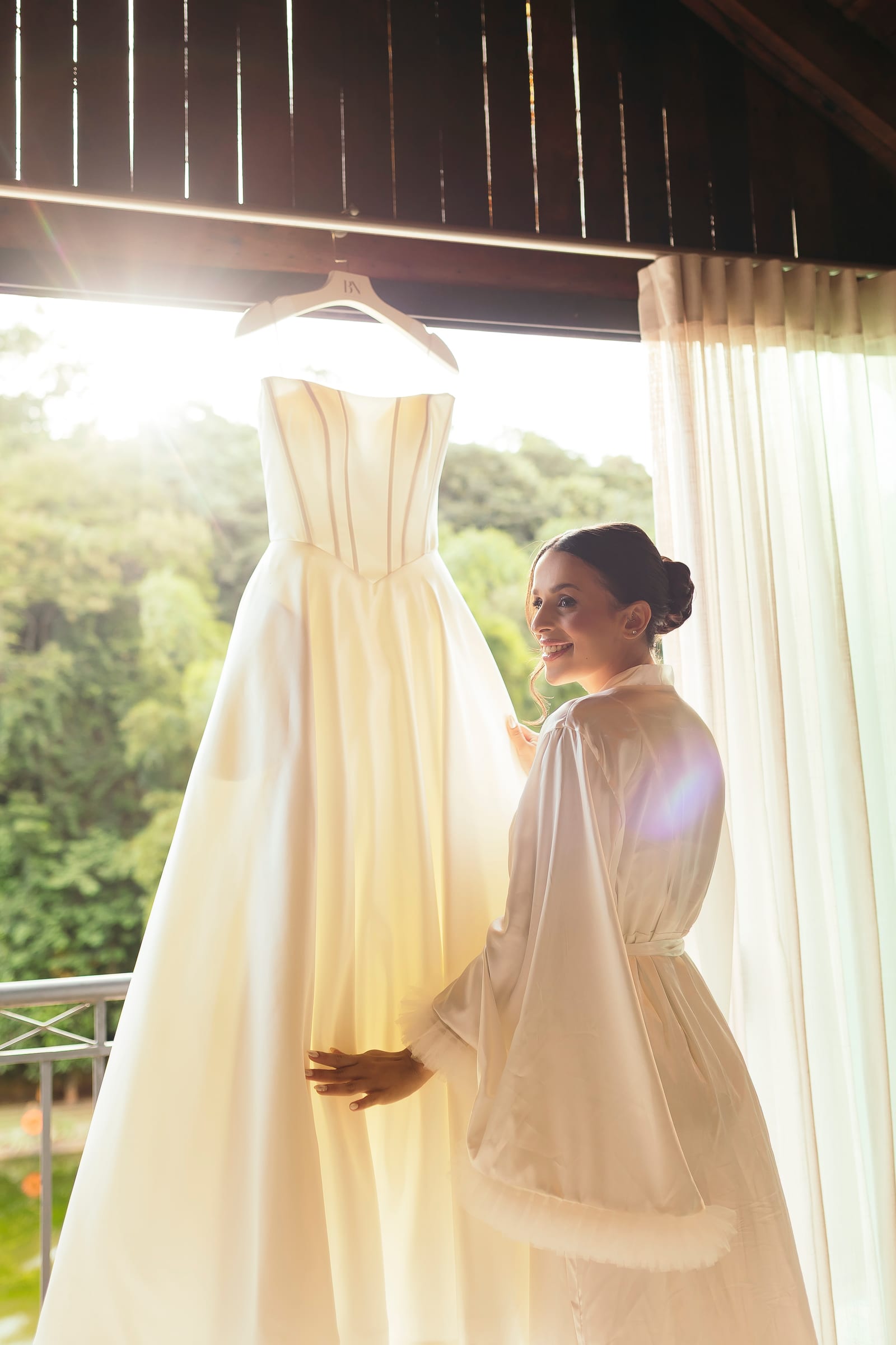 O vestido de noiva pendurado diante da janela da sala dos noivos, com a noiva de robe ao lado e a mata lá fora.