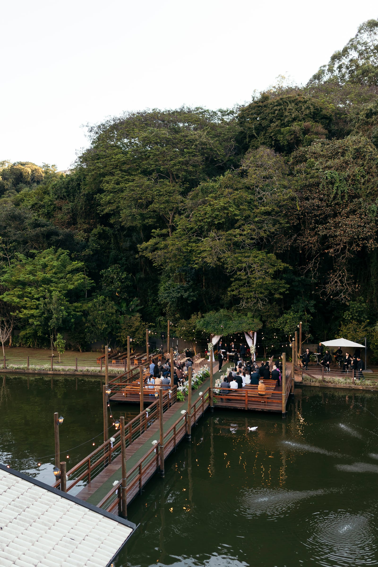 O deck sobre o lago visto do alto, com a cerimônia acontecendo e a mata fechada em volta da água.