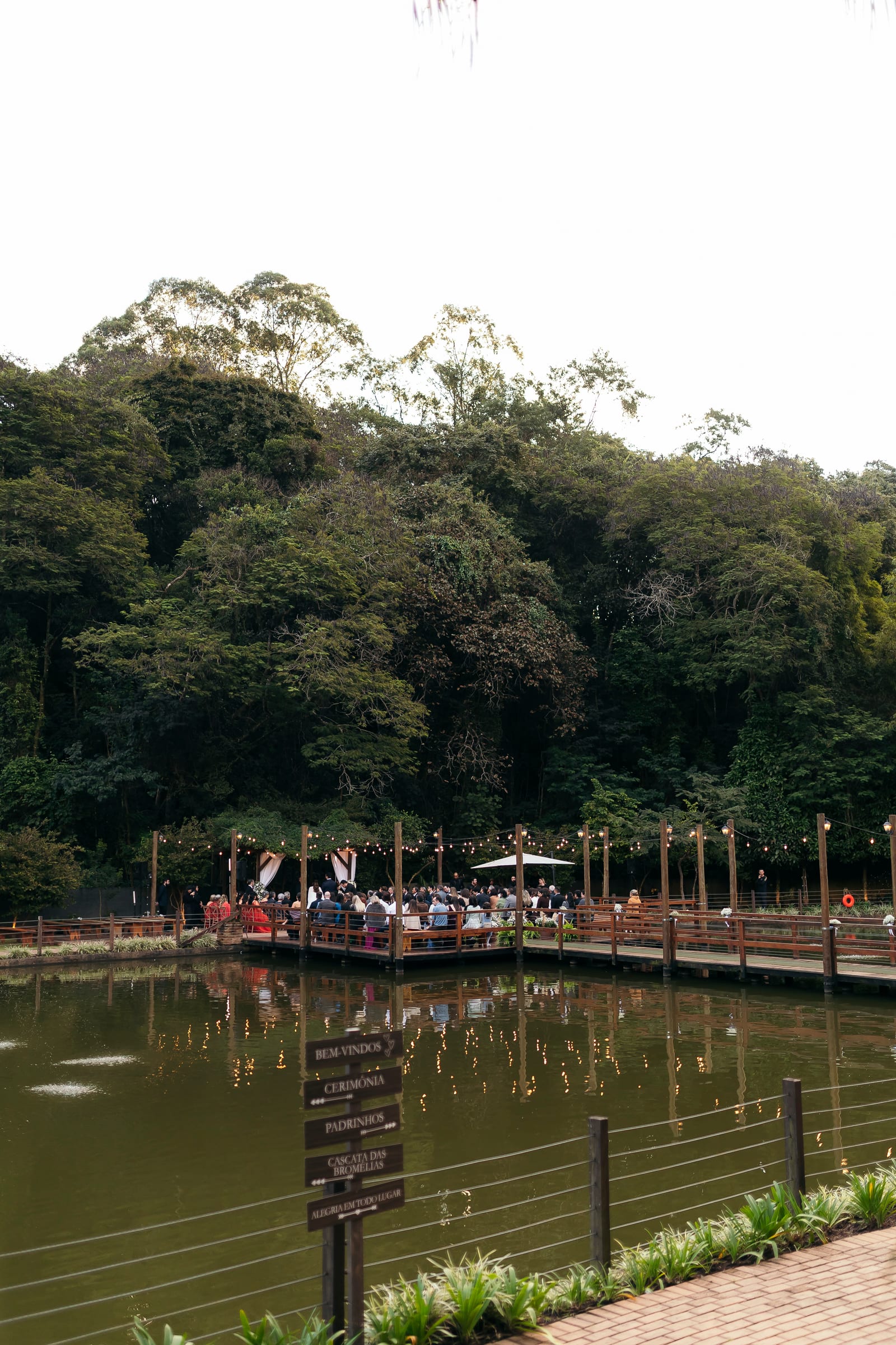 O deck sobre o lago visto da outra margem, com os convidados reunidos para a cerimônia e a placa de boas-vindas na grade.