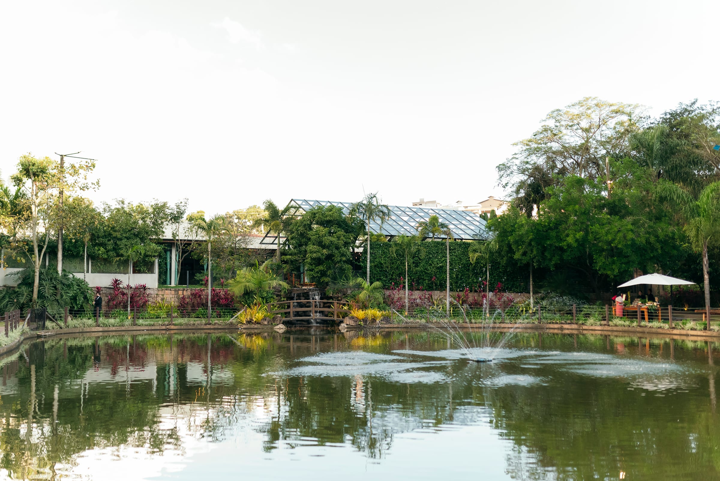 O lago do Acqua com a casa de vidro e o chafariz ao fundo, entre as árvores, sob um céu claro.
