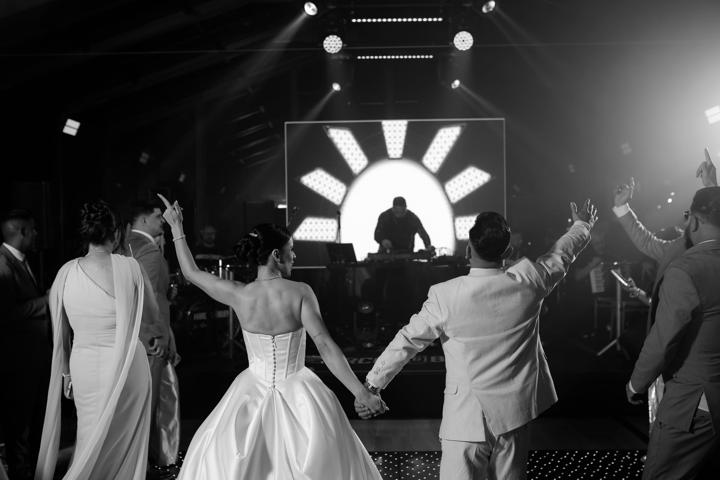 Noivos de mãos dadas diante do palco com o DJ e o painel de luzes, entre os convidados de braços erguidos, em preto e branco.