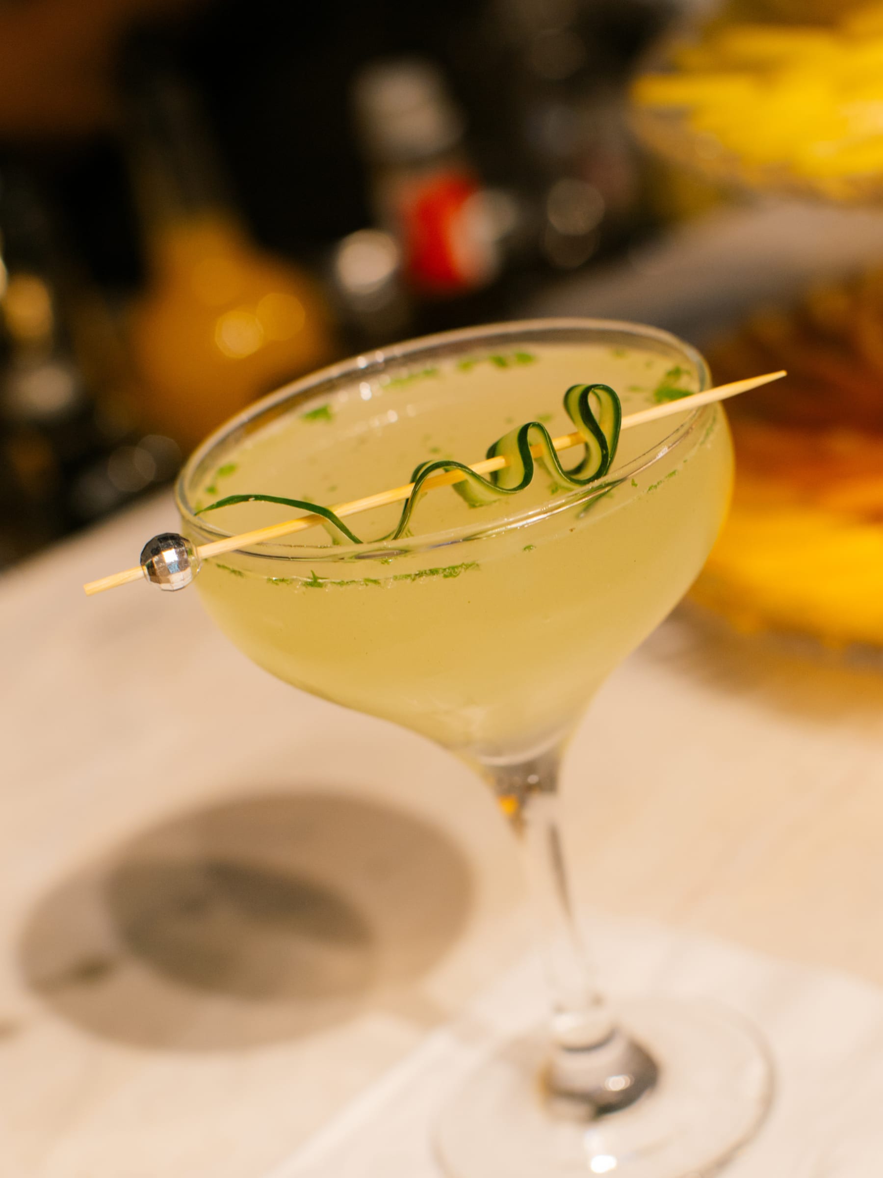 Drink verde-claro em taça coupe, com uma espiral de pepino presa num palito de bolinha espelhada.