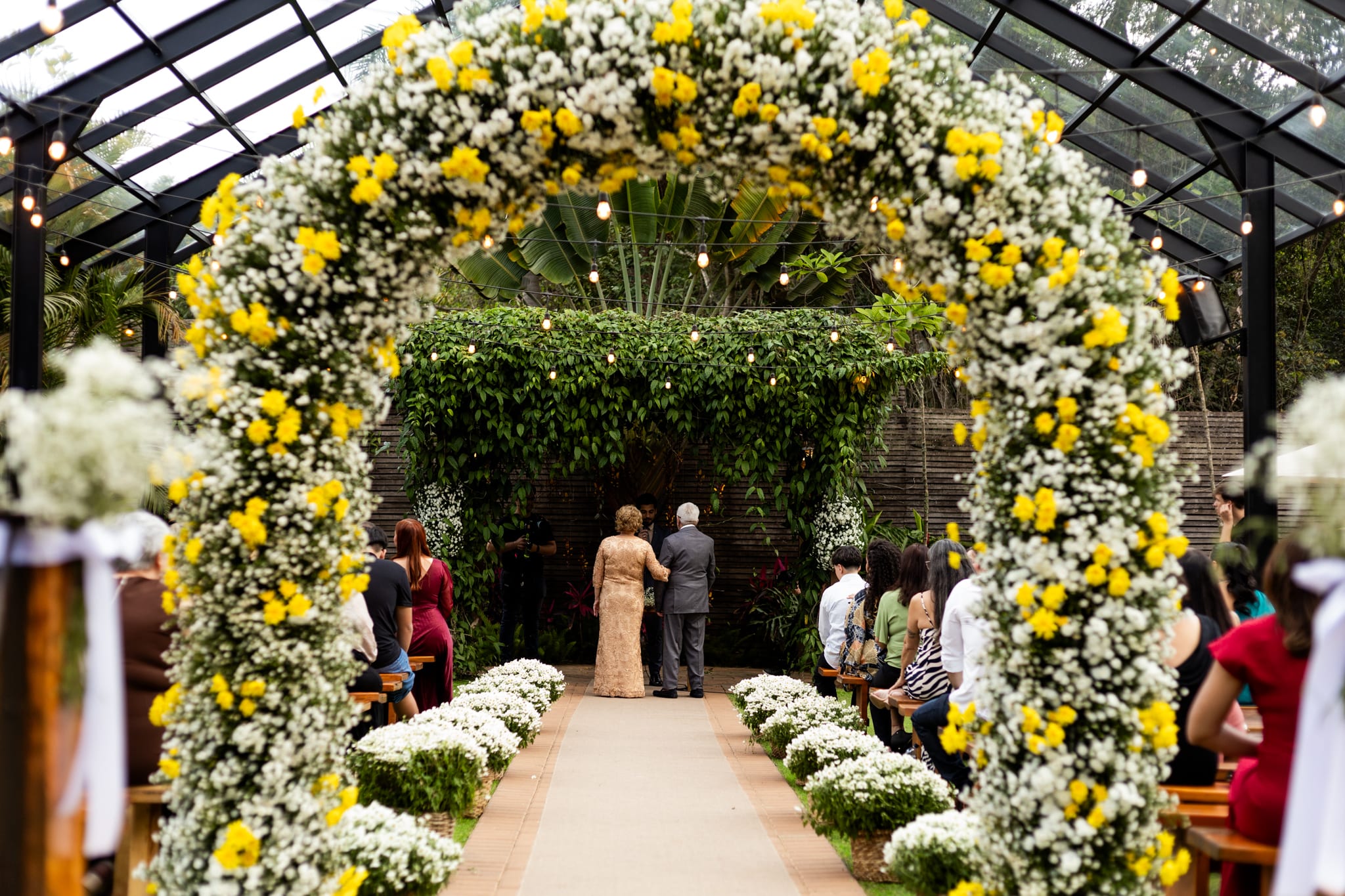 Casal renovando os votos sob um arco de flores brancas e amarelas.